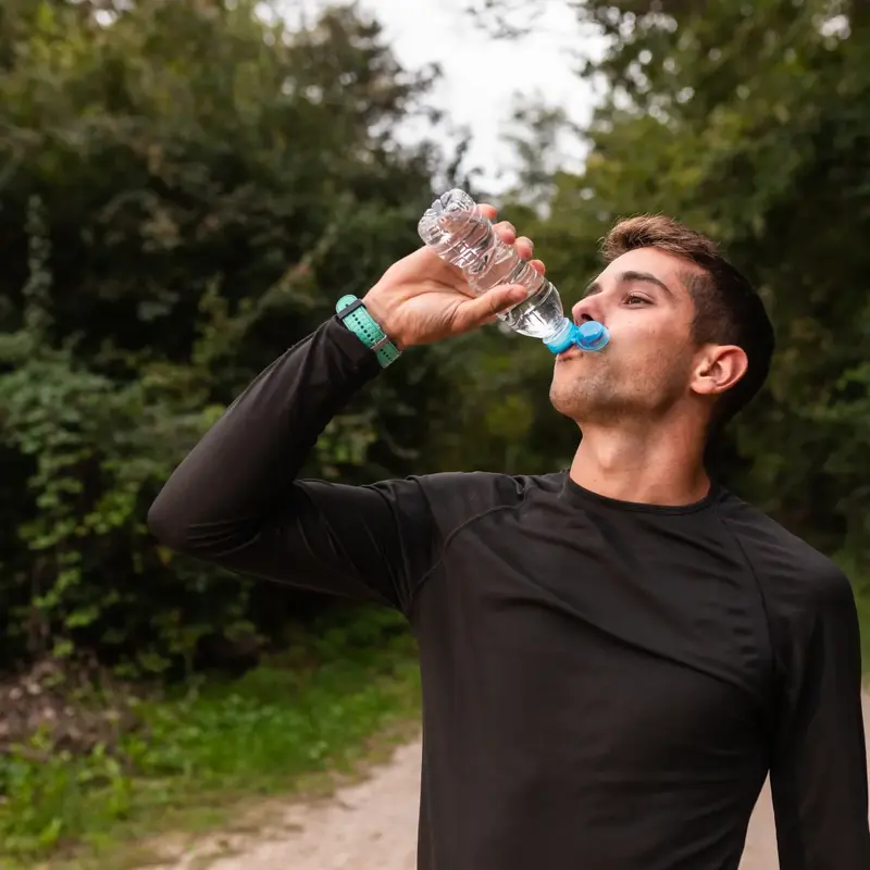 Man in black long-sleeve shirt drinks water from a plastic bottle while standing on a forest trail.