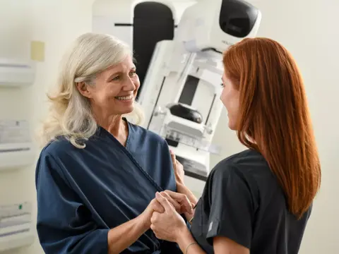 A smiling older woman and a younger woman holding hands in front of a mammogram machine.
