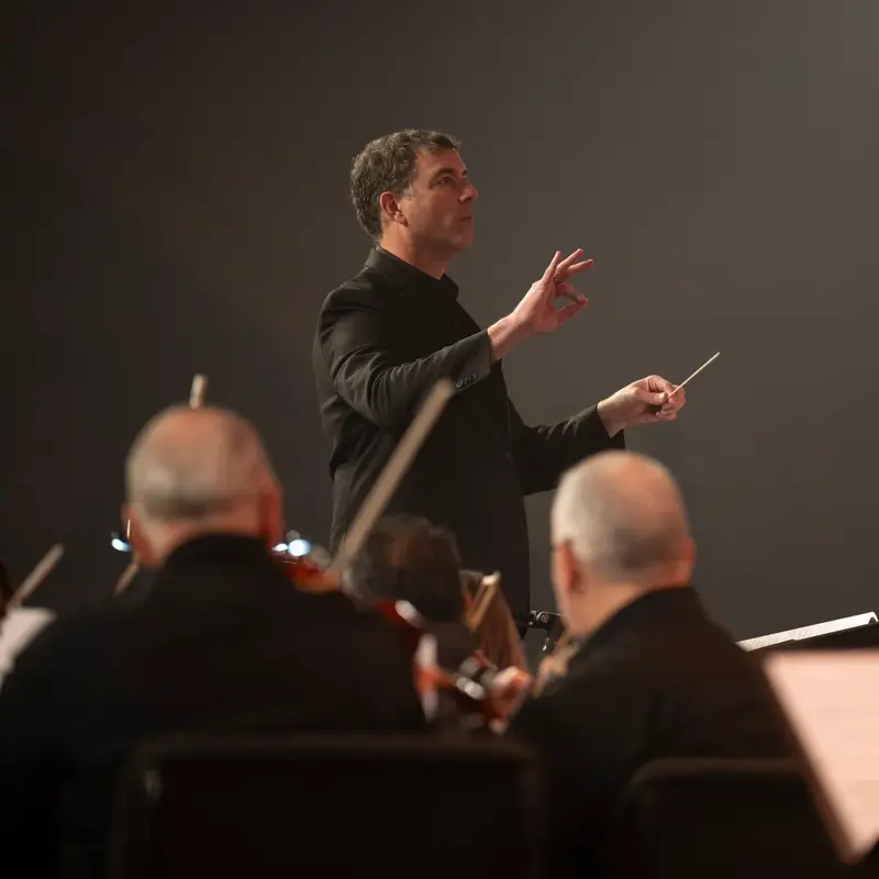 Conductor Richard Hickam leads an orchestra with a baton as musicians perform in the foreground.
