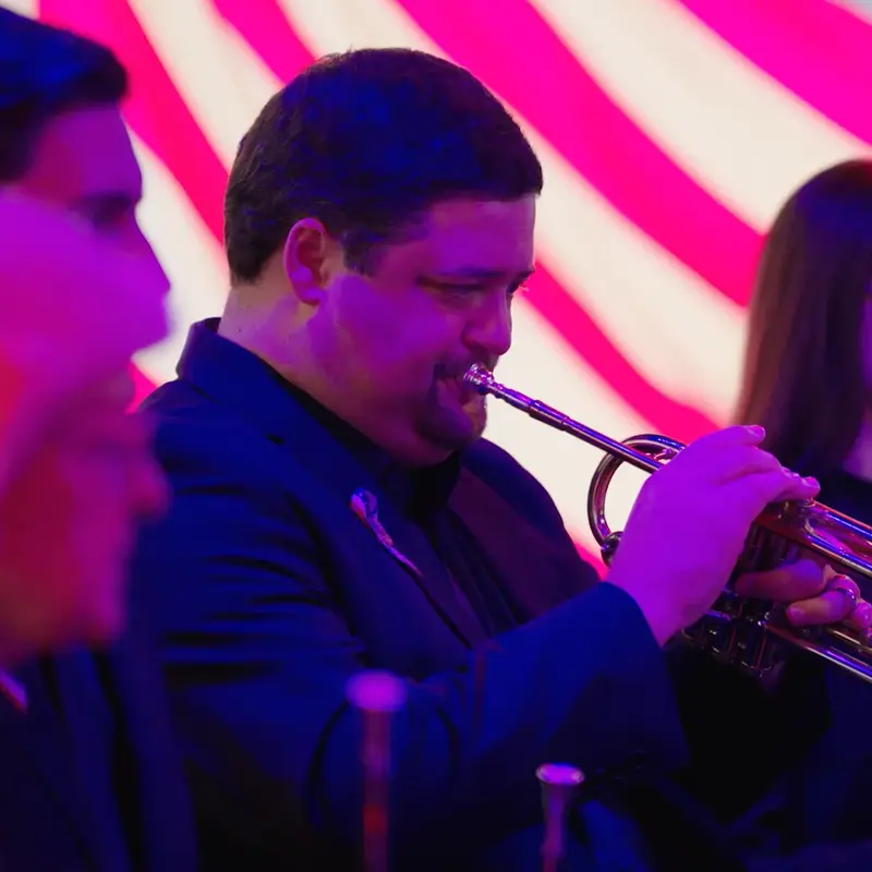 Mark Babienko plays trumpet under vivid pink and purple stage lighting, with other brass musicians behind him.