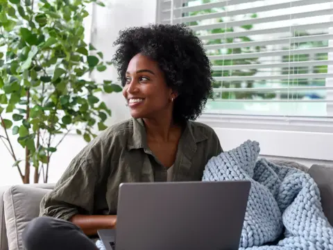 Smiling woman with laptop on couch, cozy home setting with plant and window blinds.