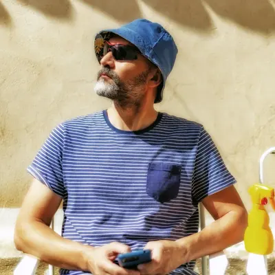 Man sitting on a beach chair in the summer sun wearing a blue striped shirt, blue hat, and sunglasses while holding a phone