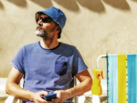 Man sitting on a beach chair in the summer sun wearing a blue striped shirt, blue hat, and sunglasses while holding a phone