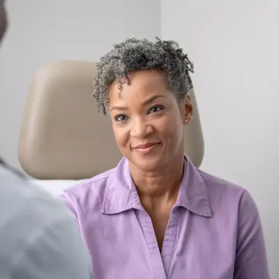 A smiling woman in a purple shirt talks with a doctor in a medical office.