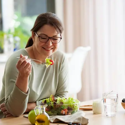 Woman smiling while holding a fork with salad at a table with bowl, fruit, and glass of water