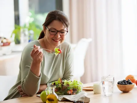 Woman smiling while holding a fork with salad at a table with bowl, fruit, and glass of water
