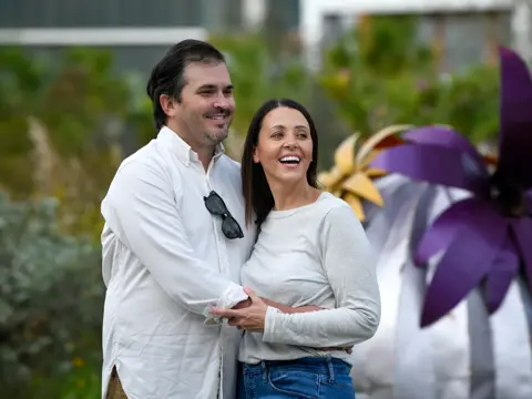 A smiling man and woman stand close together in front of a colorful garden.