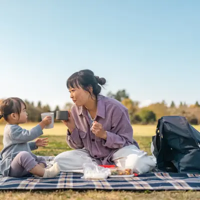 Adult and child sit on a picnic blanket outdoors, holding cups toward each other beside food and a backpack.