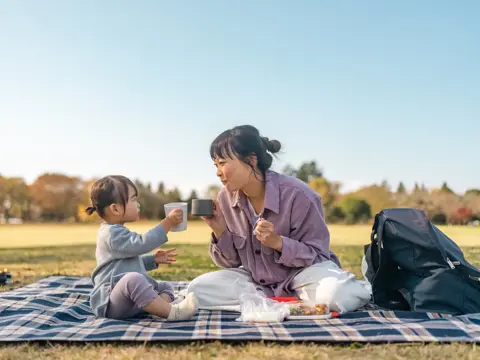 Adult and child sit on a picnic blanket outdoors, holding cups toward each other beside food and a backpack.