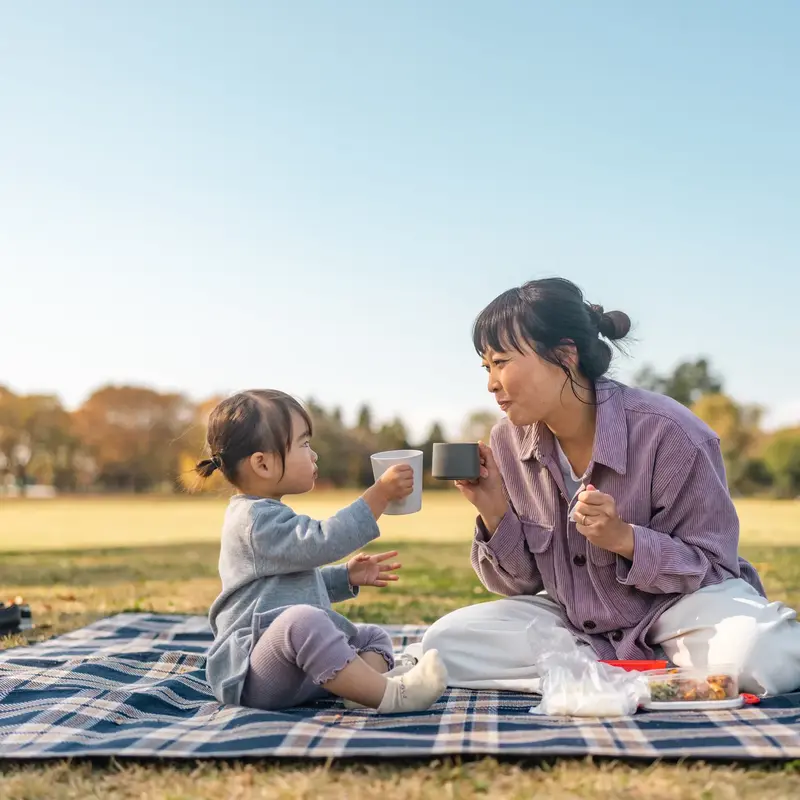 Adult and child sit on a picnic blanket outdoors, holding cups toward each other beside food and a backpack.