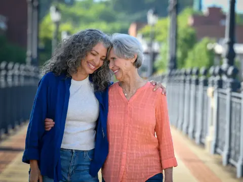 Two smiling women embracing outdoors on a sunny day, standing on a sidewalk with a blurred urban background