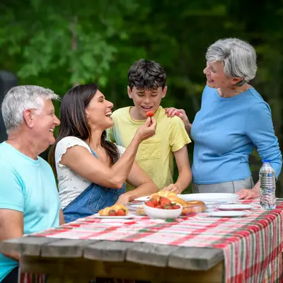 Multi-generational family of four enjoying a picnic outdoors, with a mother offering a strawberry to her young son while grandparents smile at a table with a red checkered tablecloth