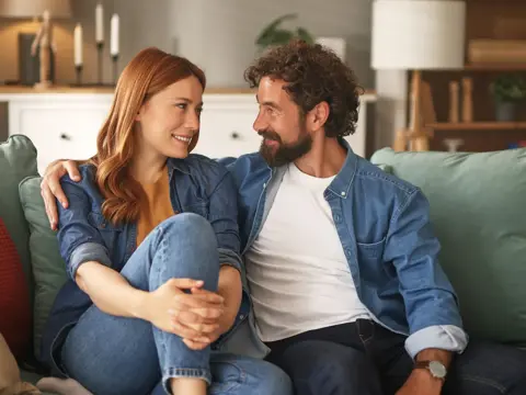 A smiling couple sitting close together on a couch in a warmly lit living room, engaged in an intimate conversation