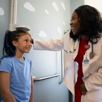 A smiling doctor in a white coat and red shirt examines a young girl with blue hair.