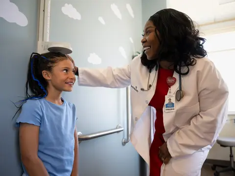 A smiling doctor in a white coat and red shirt examines a young girl with blue hair.