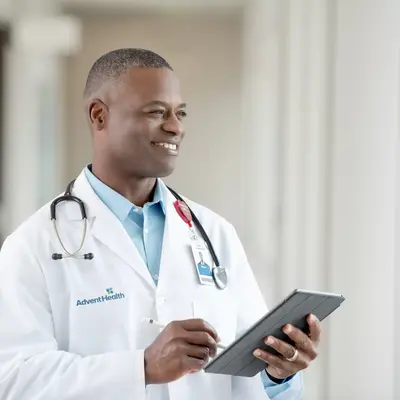 Smiling doctor in white coat with stethoscope holding a tablet, standing in a hospital corridor.