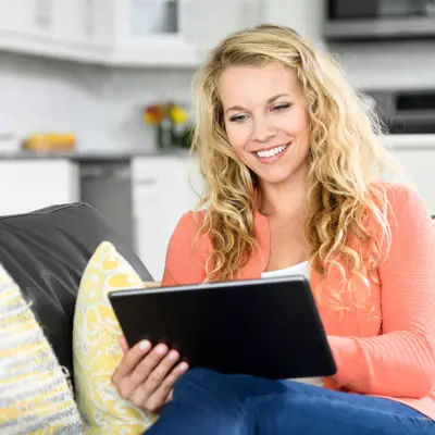 A smiling woman with blonde hair sits on a couch, holding a tablet, in a cozy living room.