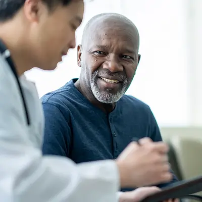 Doctor in white coat consulting with an older male patient in a blue shirt, reviewing information together in a bright clinical setting