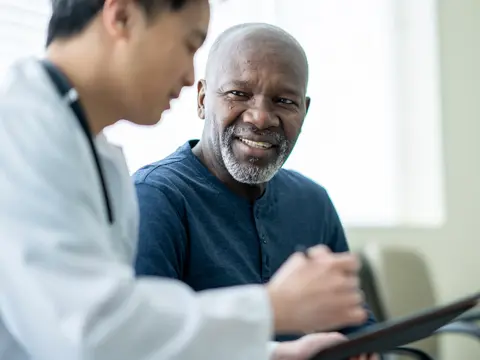 Doctor in white coat consulting with an older male patient in a blue shirt, reviewing information together in a bright clinical setting