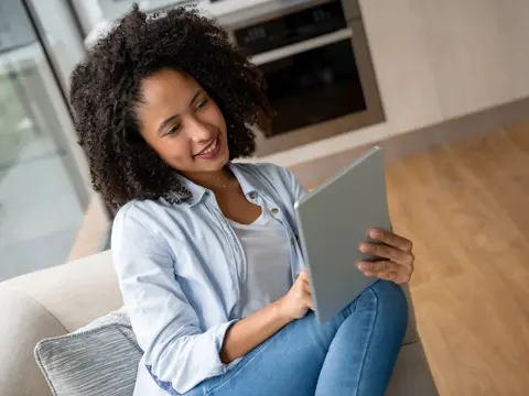 Person with curly hair sits on a couch, smiling while using a tablet in a bright living room.