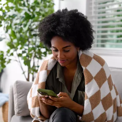 A woman wrapped in a blanket, using her phone while sitting on a couch.