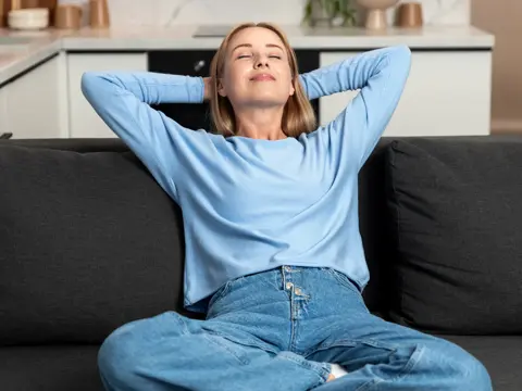 Woman in a light blue long-sleeve shirt sitting on a dark gray couch with her eyes closed and hands behind her head, relaxing in a bright modern kitchen setting.