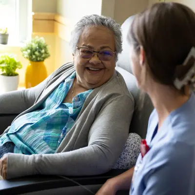 Elderly woman with glasses smiling while sitting in a chair and talking to a nurse.