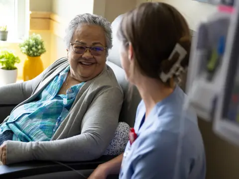 Elderly woman with glasses smiling while sitting in a chair and talking to a nurse.