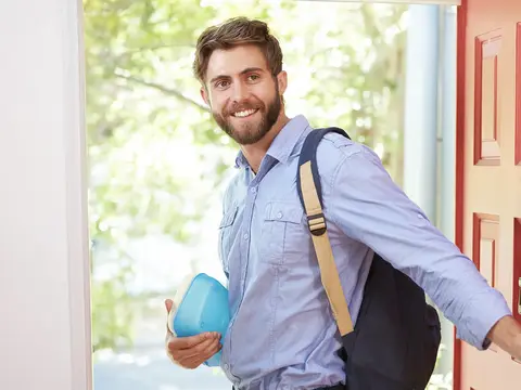 Smiling man with a backpack and blue container opens a red front door.