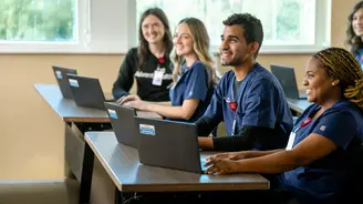 Group of nursing residents in navy scrubs smiling and working on laptops during a training session in a bright classroom with windows