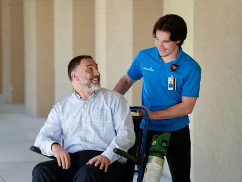 A smiling man in a wheelchair is being assisted by a healthcare worker in a blue shirt.