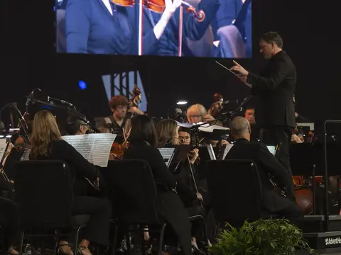 Orchestra performs onstage as a conductor leads musicians beneath a large screen showing violinists.