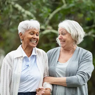 Two multiracial senior women conversing, walking together outdoors in a back yard, with trees behind them.