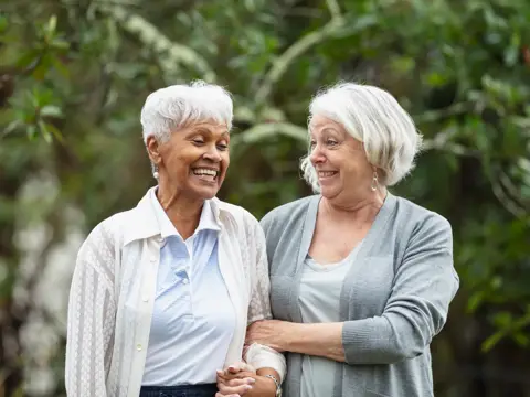 Two multiracial senior women conversing, walking together outdoors in a back yard, with trees behind them.