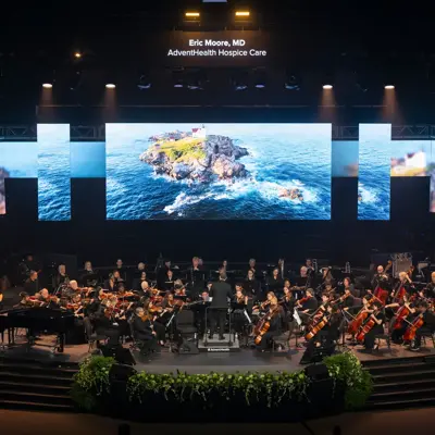 Orchestra performing on stage with large screens displaying scenic images and text 'Eric Moore, MD AdventHealth Hospice Care'.