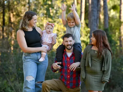 Family of five smiling in a forest, with a man holding a young boy on his shoulders.
