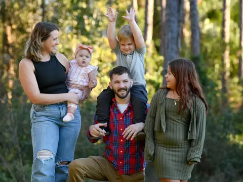 Family of five smiling in a forest, with a man holding a young boy on his shoulders.