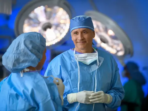 Surgeon in blue scrubs and cap smiling at another surgeon in an operating room.