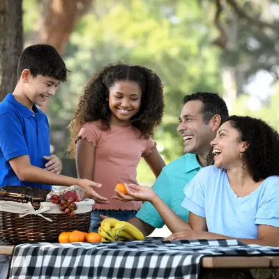 Family of four sharing a picnic with fruit and bananas on a sunny day.
