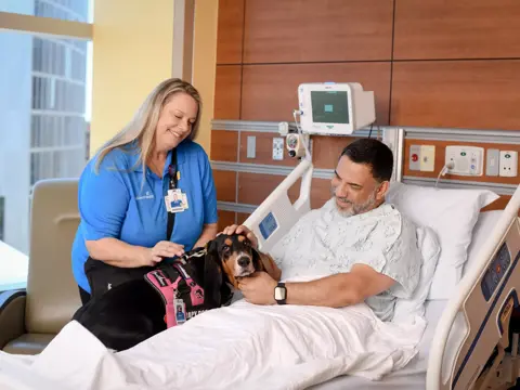 A smiling patient in a hospital bed with a therapy dog and a nurse.