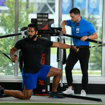 A man performs a workout with resistance bands while a trainer observes, both in a gym setting.