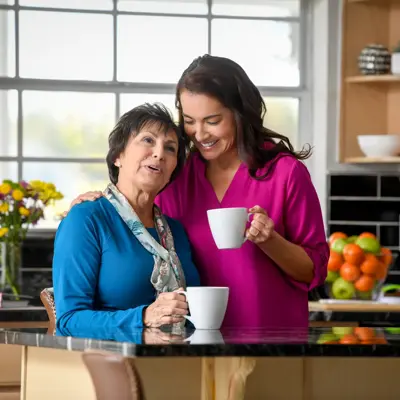 Two women in a kitchen, one holding a mug, smiling and engaged in conversation.