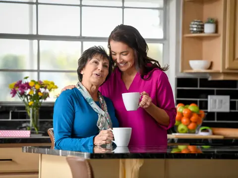 Two women in a kitchen, one holding a mug, smiling and engaged in conversation.