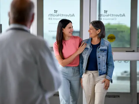 Two smiling women stand together outside a glass door, with a doctor in the background. AdventHealth logo visible.