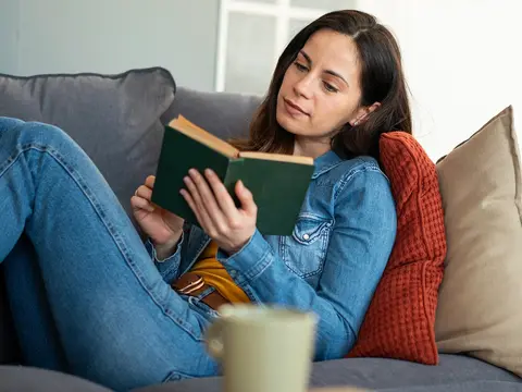 Woman in denim jacket and orange sweater reading a green book while relaxing on a gray couch with pillows