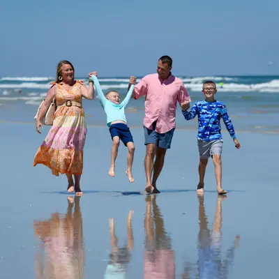 Family of four holding hands and walking on the beach with ocean waves in the background.