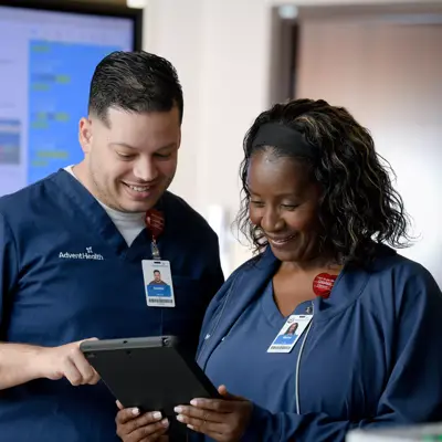 Two AdventHealth employees in scrubs, smiling and looking at a tablet together.