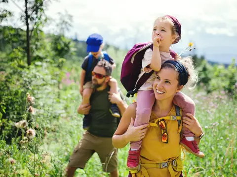 Family of four hiking through a vibrant green meadow with mountains in the background, parents carrying young children on their shoulders while holding wildflowers