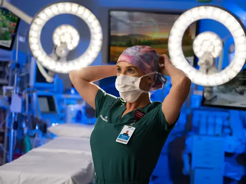 A nurse in green scrubs and a mask stands in an operating room with surgical lights.