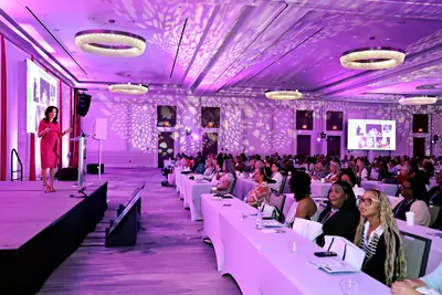 Speaker presenting on stage to a large audience during a conference in a ballroom illuminated with purple lighting.